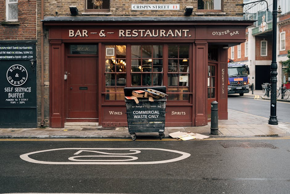 A black wheeled commercial waste skip with a hinged lid is placed on the pavement in front of a restaurant with a brown wooden exterior and large glass windows. The skip has white text on the front reading 'COMMERCIAL WASTE ONLY' and is filled with various unwanted items, including flattened cardboard boxes and loose paper. Nearby, a small piece of cardboard rests on the ground beside the skip. The scene is set on a street with a painted white bus stop symbol on the asphalt directly in front of the waste container. To the left, there is a blackboard with white chalk writing advertising a self-service buffet, and a black bollard is positioned on the right side of the skip, securing the edge of the sidewalk. A black lamppost and a parked large truck are visible in the background, along with additional bicycles attached to a bike rack on the sidewalk. The overall setting suggests an urban environment where independent commercial waste collection might be conducted by local waste management services such as Rubbish Clearance Bayswater, ensuring tidy disposal of business-related refuse in a public area.