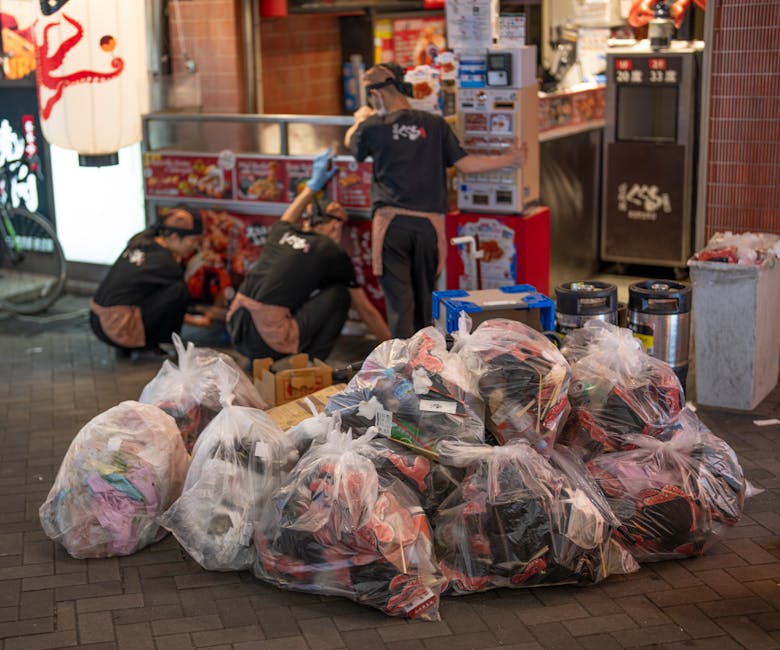 A black wheeled commercial waste skip with a hinged lid is placed on the pavement in front of a restaurant with a brown wooden exterior and large glass windows. The skip has white text on the front reading 'COMMERCIAL WASTE ONLY' and is filled with various unwanted items, including flattened cardboard boxes and loose paper. Nearby, a small piece of cardboard rests on the ground beside the skip. The scene is set on a street with a painted white bus stop symbol on the asphalt directly in front of the waste container. To the left, there is a blackboard with white chalk writing advertising a self-service buffet, and a black bollard is positioned on the right side of the skip, securing the edge of the sidewalk. A black lamppost and a parked large truck are visible in the background, along with additional bicycles attached to a bike rack on the sidewalk. The overall setting suggests an urban environment where independent commercial waste collection might be conducted by local waste management services such as Rubbish Clearance Bayswater, ensuring tidy disposal of business-related refuse in a public area.