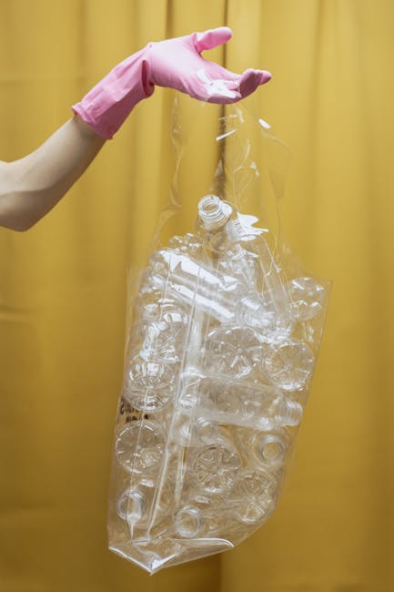 A person wearing a pink rubber glove is holding a large, clear plastic bag filled with numerous empty plastic bottles, most of which are transparent and some with visible caps. The plastic bottles vary in size and shape, with some having ridged sides and others smooth, and are loosely stacked inside the bag. The background consists of a plain, muted yellow curtain or wall, creating a neutral environment that emphasizes the plastic waste collection. The scene suggests an effort towards recycling or waste separation, aligning with private waste handling and rubbish clearance practices offered by Rubbish Clearance Bayswater, particularly in the context of household rubbish removal on Porchester Road and across the W2 postcode area. The overall composition highlights the focus on plastic waste, a common component of household rubbish, and reflects the importance of proper disposal and recycling strategies in contemporary waste management.