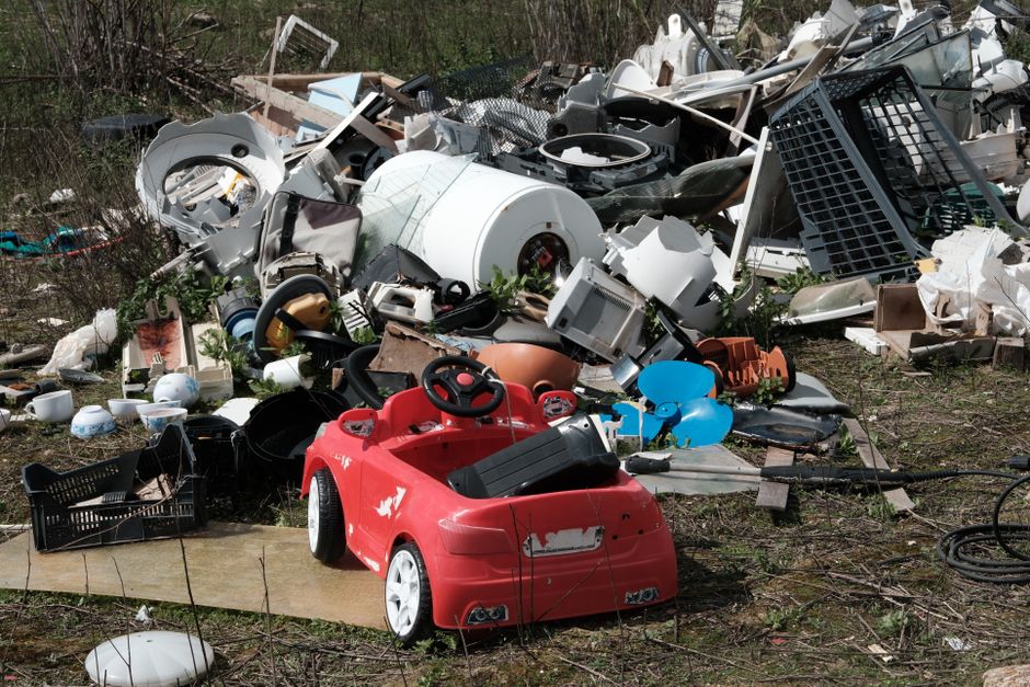 An outdoor scene showing a large pile of discarded household and electronic waste items scattered on bare ground with patches of grass and sparse vegetation in the background. In the foreground, there is a small, red plastic toy car with a slightly weathered finish, featuring a steering wheel and black seat, positioned beside a black plastic crate and surrounded by various smaller debris. The main pile contains an array of broken appliances, including a white washing machine drum, various metal and plastic panels, circuit boards, and other electronic components, some with visible textures and finishes such as glossy white surfaces, matte black casings, and rusted metal parts. There are also plastic containers, pipes, and household items mixed in, with some partially buried or leaning against each other, creating a chaotic arrangement typical of on-site rubbish clearance sites. Natural lighting illuminates the scene, emphasizing the textures and materials of the discarded objects, which are related to private waste disposal and alternative rubbish removal methods often handled by professional services like Rubbish Clearance Bayswater.