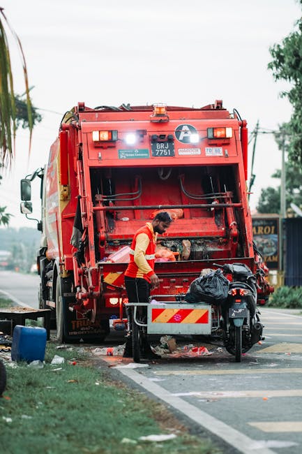 A street scene shows a worker in a red and yellow uniform operating a large red waste collection vehicle, positioned on the side of the road with its rear hatch open, revealing mechanical components and compartments for rubbish. The worker faces downward, handling black plastic waste bags that are placed on a metal platform attached to a motorcycle parked nearby. The motorcycle, with a black seat and shades of grey, is situated close to the waste vehicle, and a traffic cone is visible behind the motorcycle. The background includes a cloudy sky, some trees with green foliage, and a building with a mural or signage on the side, indicating an urban environment. The pavement around the waste collection scene shows some scattered debris and is bordered by a grassy verge. The scene is well-lit, suggesting daytime, and emphasizes the process of independent waste collection, aligning with private rubbish removal services offered by companies such as Rubbish Clearance Bayswater.